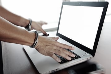 Businessman hand in handcuffs at wooden desk with laptop computer and digital tablet and stylus pen and smart phone as Cyber-Crime concept